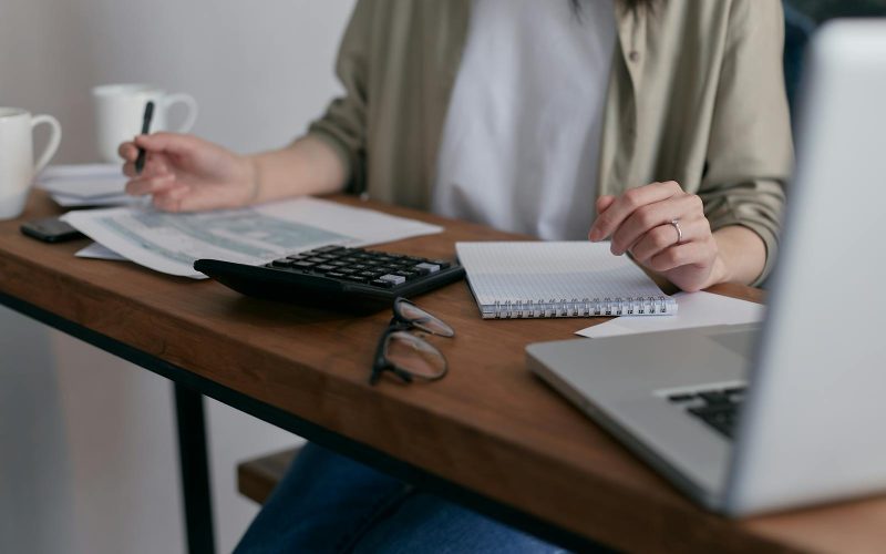 A woman manages finances at home, using a laptop and calculator on a wooden desk.